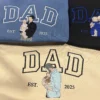 Indigo blue, black and sand sweatshirts with outline DAD letters and embroidered dad-and-baby portraits, EST 2025