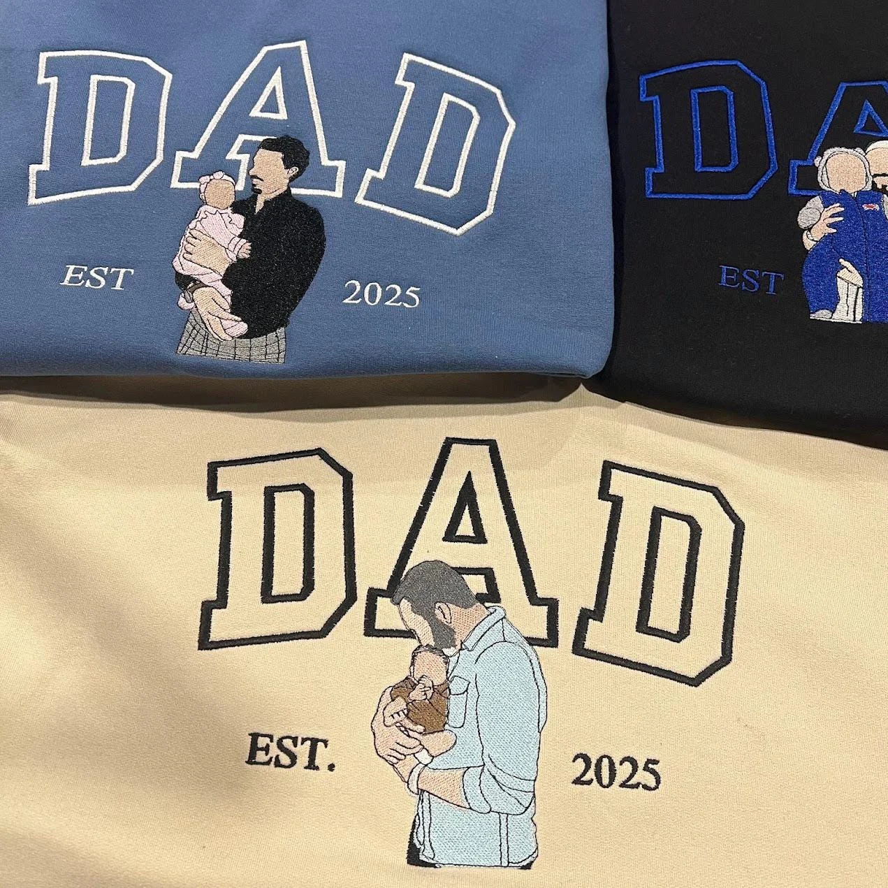 Indigo blue, black and sand sweatshirts with outline DAD letters and embroidered dad-and-baby portraits, EST 2025