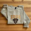 Sand crewneck laid on a wooden floor with an embroidered chocolate Labrador under the name ZEUS in varsity letters