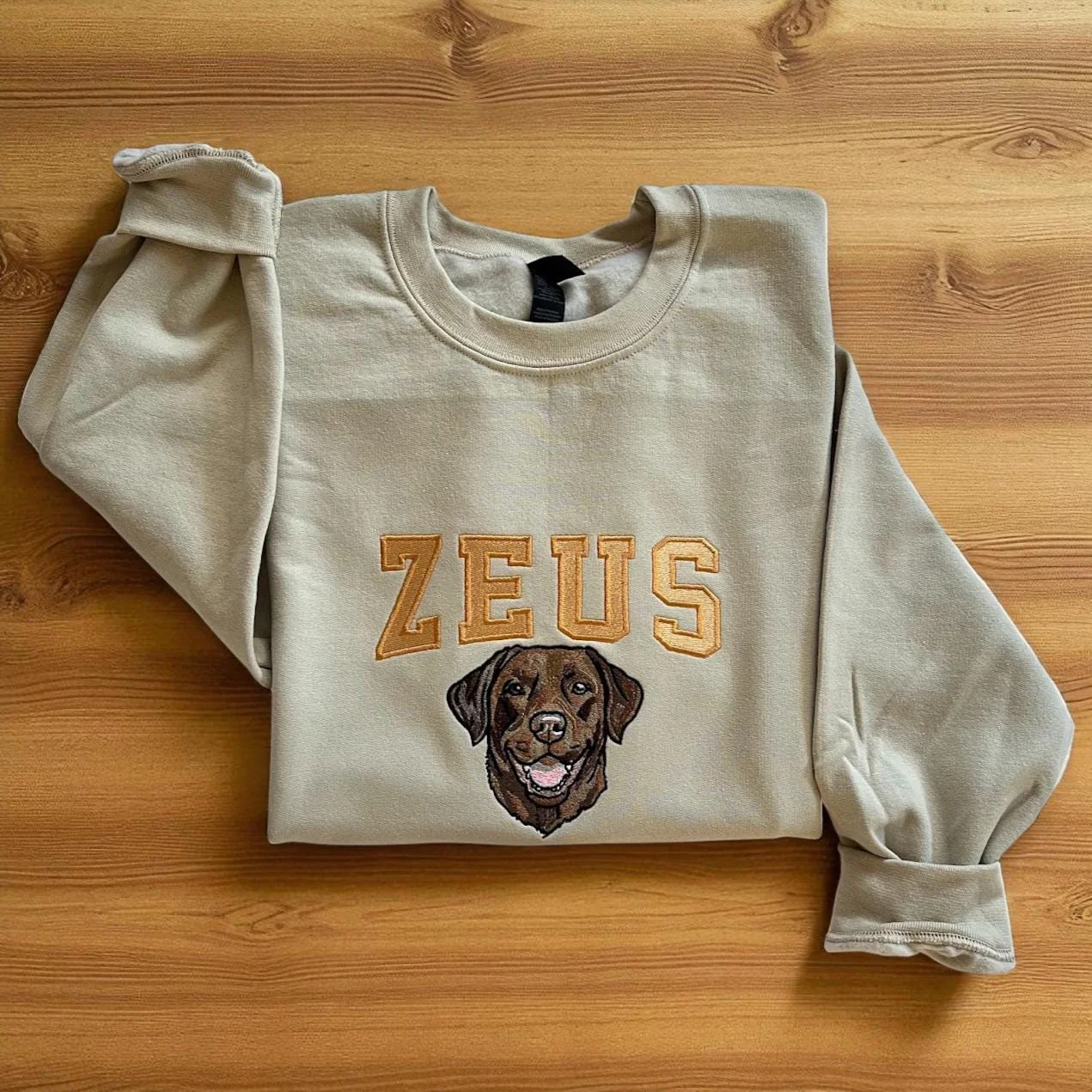 Sand crewneck laid on a wooden floor with an embroidered chocolate Labrador under the name ZEUS in varsity letters