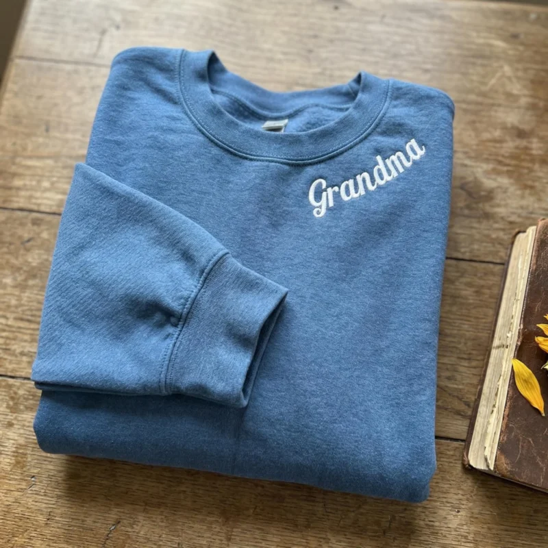 Folded heathered blue sweatshirt with Grandma embroidered in white script beside the neckline, on a wooden table