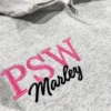 Close-up of an ash quarter-zip with PSW in pink serif capitals and Marley in black script across them