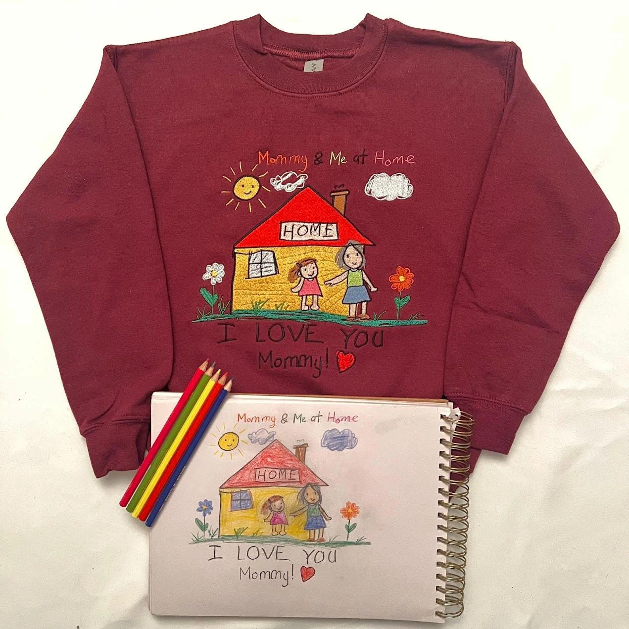 Maroon crewneck laid flat with the embroidered child’s house drawing, colouring pencils and the original sketchbook