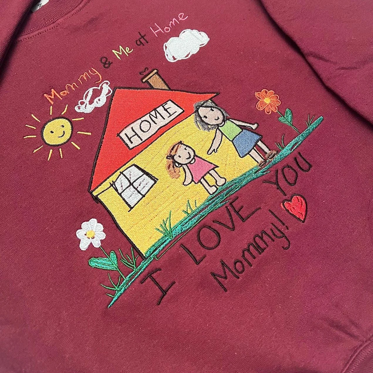 Angled close-up of I LOVE YOU Mommy and a red heart under an embroidered crayon-style house and family