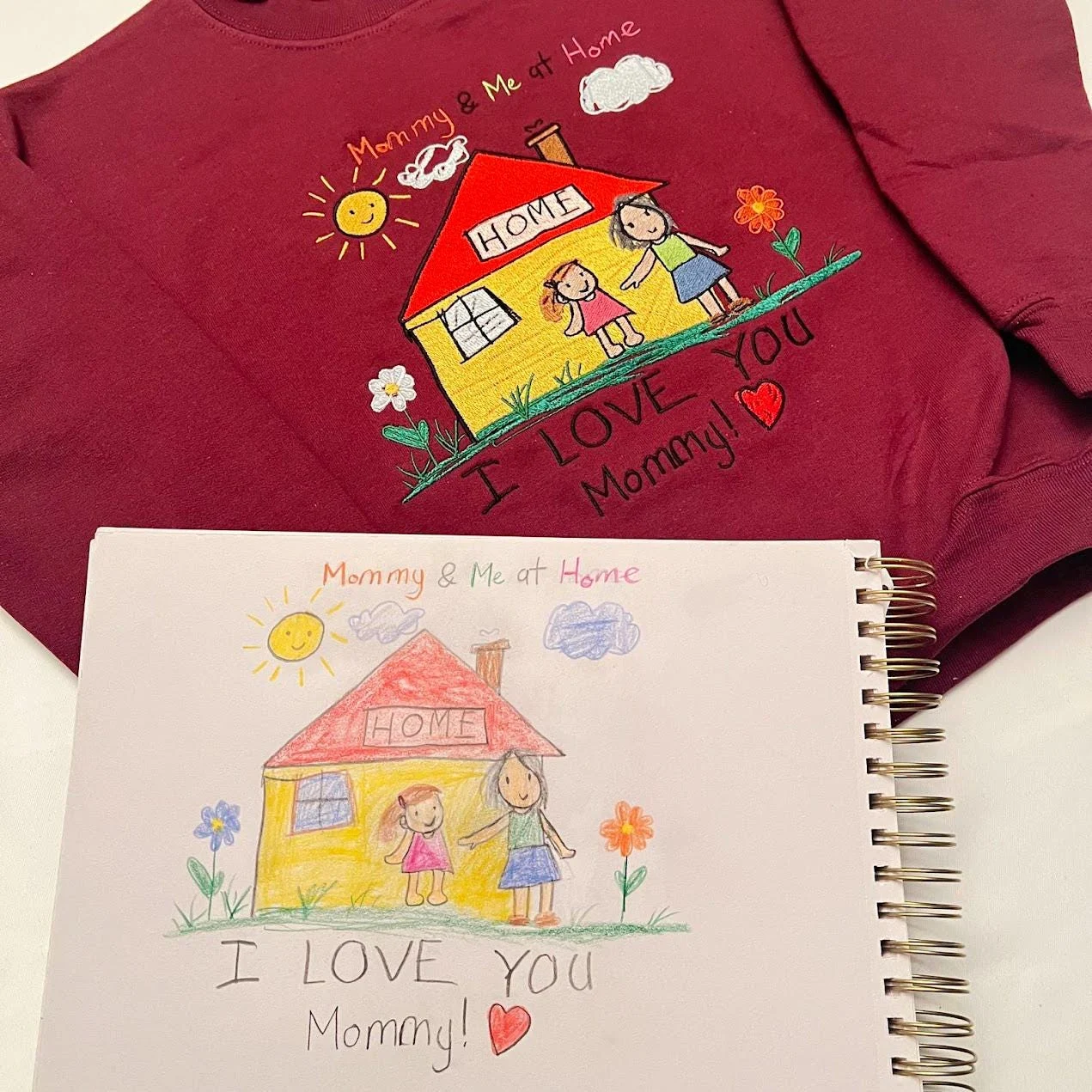 Embroidered child’s drawing Mommy and Me at Home on a maroon sweatshirt next to the sketchbook drawing it copies