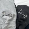Close-up of the name Aminah with buzzing bees embroidered on the cuffs of grey and black sweatshirts