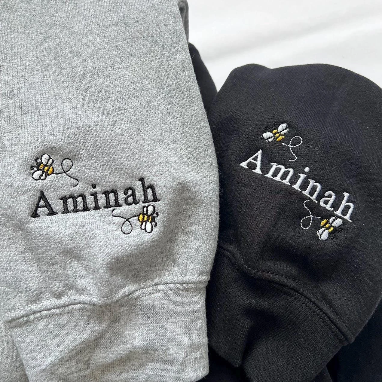 Close-up of the name Aminah with buzzing bees embroidered on the cuffs of grey and black sweatshirts