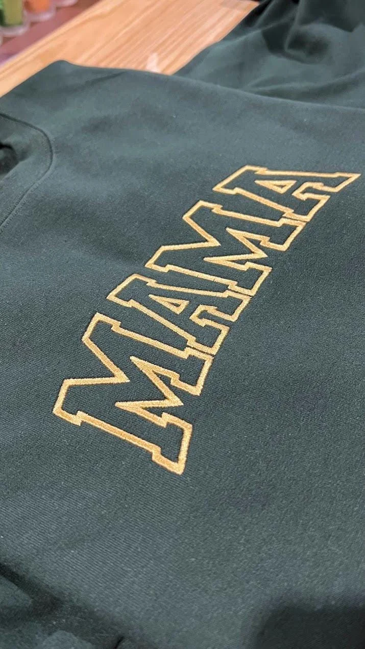 Close-up of arched MAMA varsity lettering with a gold outline embroidered on a forest green sweatshirt