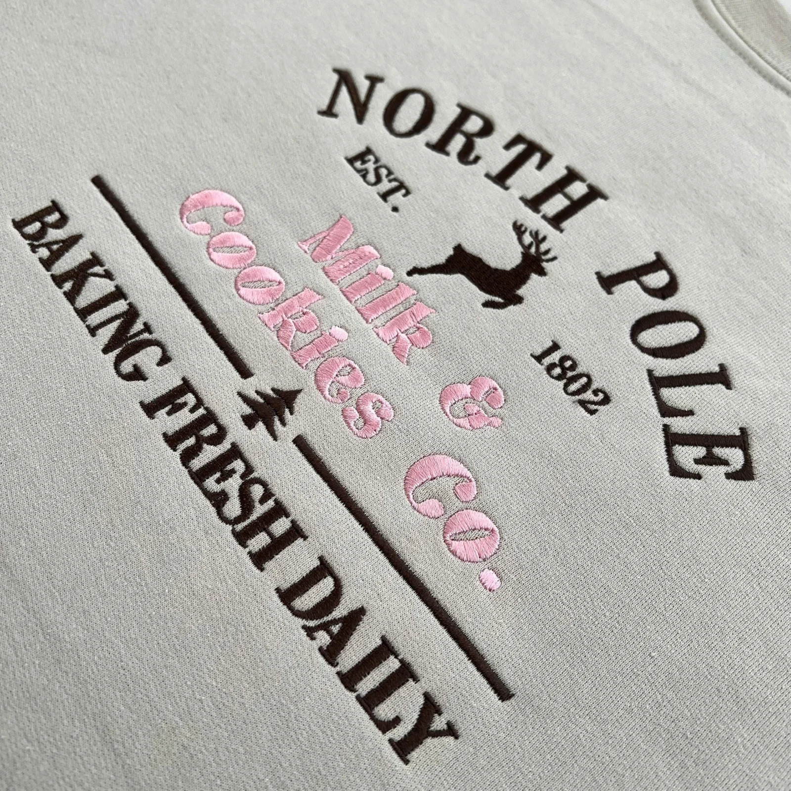 Diagonal close-up of the reindeer bakery sign embroidery in dark brown and pink thread on a sand sweatshirt