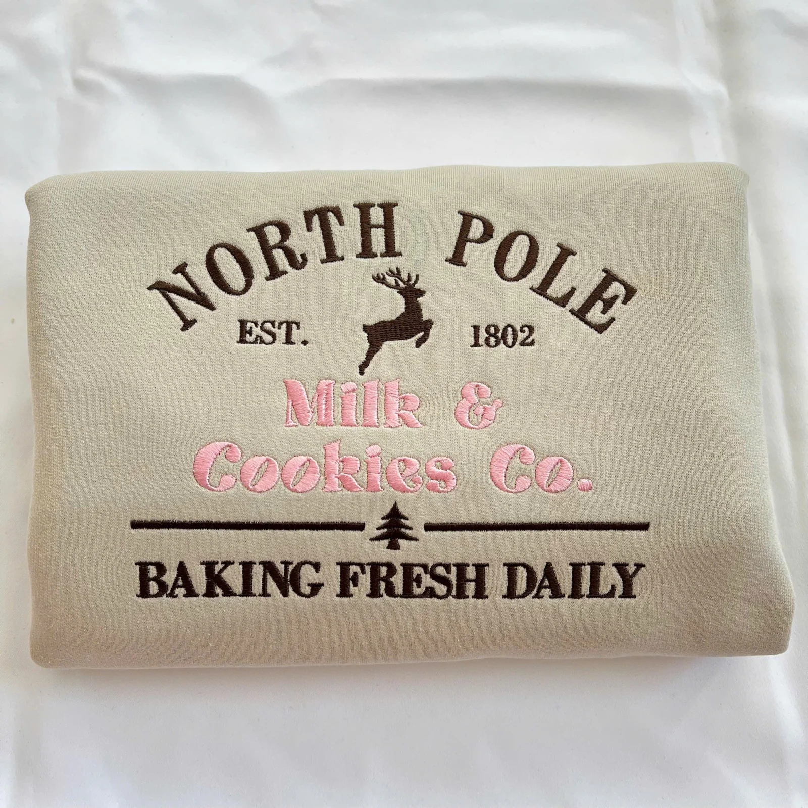 Folded sand sweatshirt with NORTH POLE in brown, a reindeer, EST. 1802 and Milk & Cookies Co. in pink