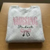 Folded heather grey sweatshirt with NURSING in pink capitals, Student in black script and a pink bow with stethoscope