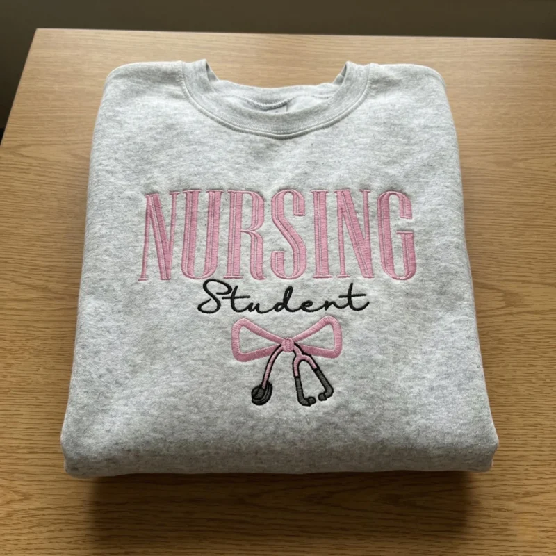 Folded heather grey sweatshirt with NURSING in pink capitals, Student in black script and a pink bow with stethoscope
