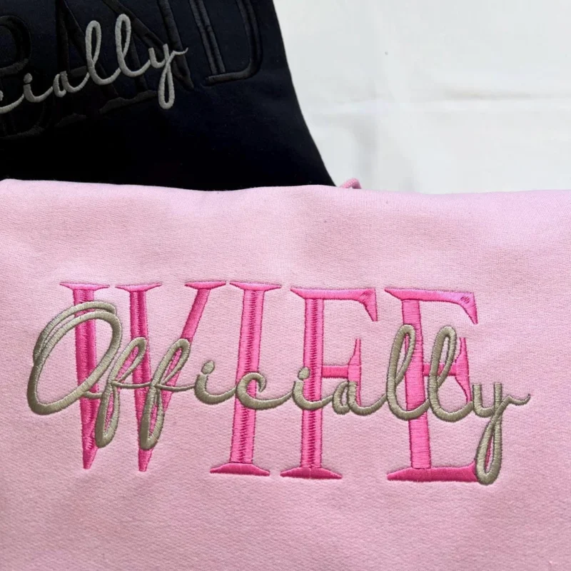 Light pink hoodie with WIFE in pink serif capitals and Officially in grey script across it, black hoodie behind