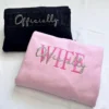 Folded black HUSBAND and pink WIFE hoodies, each with Officially stitched in script across the capital letters