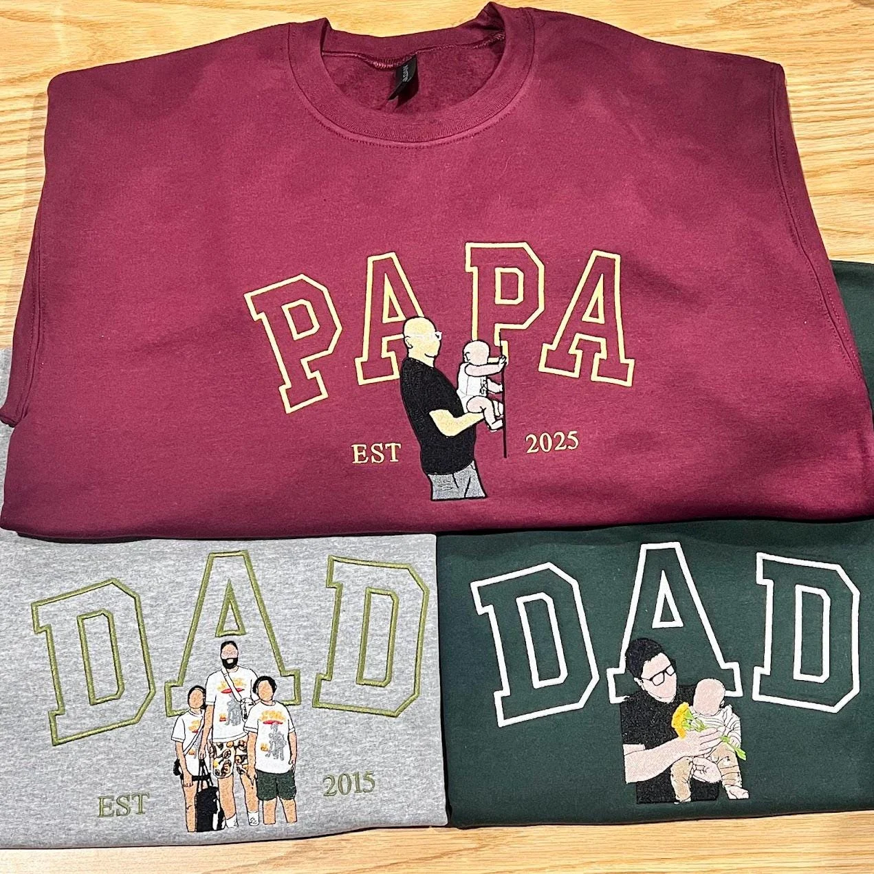 Maroon PAPA sweatshirt with a dad lifting a baby, grey DAD with a family of four, and green DAD with dad and baby