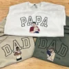 White sweatshirt with outline PAPA and sand and green hoodies with DAD, each with a stitched dad holding a child