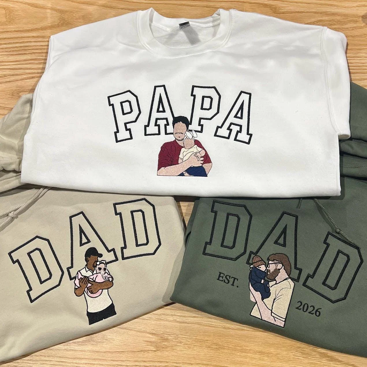 White sweatshirt with outline PAPA and sand and green hoodies with DAD, each with a stitched dad holding a child