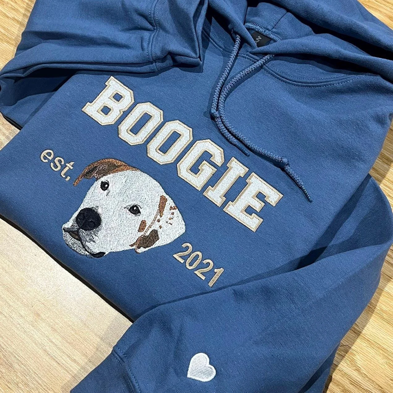 Indigo blue hoodie with a dog portrait, varsity name and est. 2021, plus a small white heart on the sleeve cuff