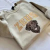 Sand sweatshirt with ZEUS in caramel varsity letters and a thread-painted chocolate Labrador face beside it