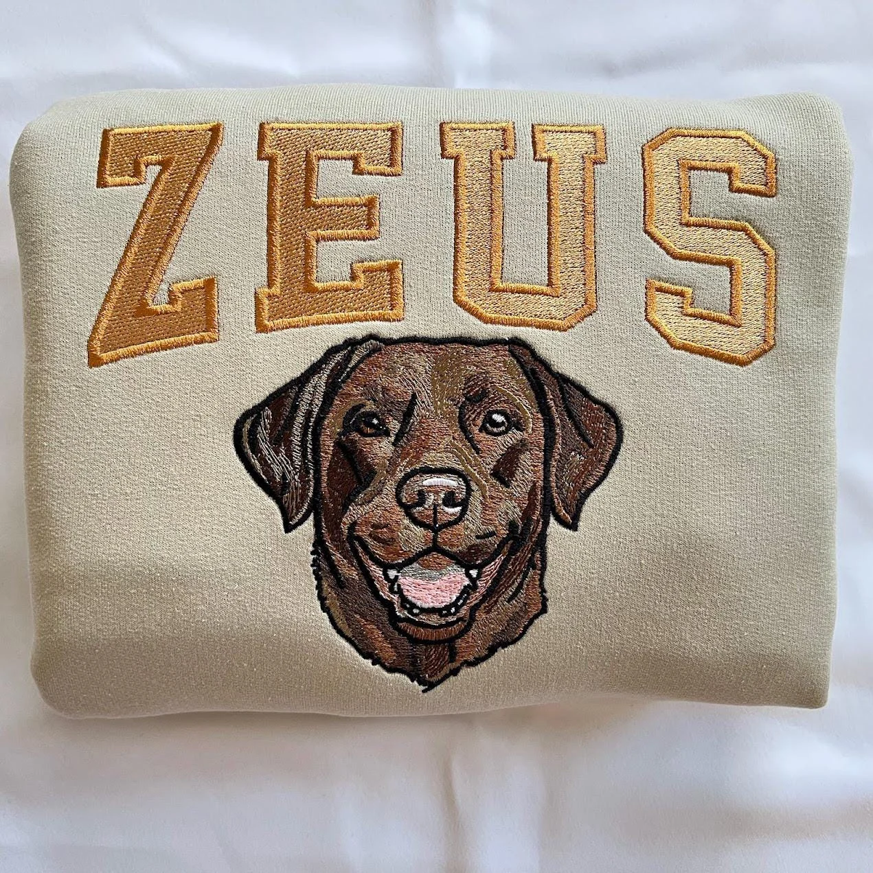 Folded sand sweatshirt with large ZEUS varsity letters above a detailed chocolate Labrador face embroidery