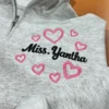 Ash quarter-zip with Miss. Yantha in black script surrounded by pink outline hearts on the chest
