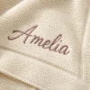 Close-up of Amelia embroidered in mauve script on an ivory moss-stitch knit baby blanket near its border