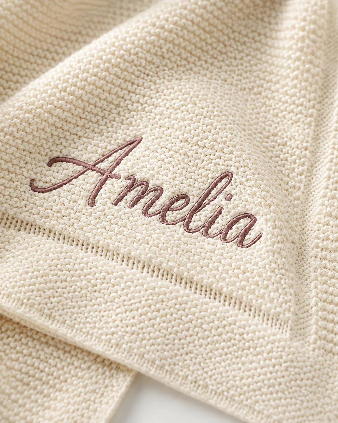 Close-up of Amelia embroidered in mauve script on an ivory moss-stitch knit baby blanket near its border