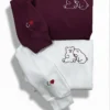 Maroon sweatshirt above a white one, each embroidered with two hugging bears and an initial and heart on the sleeve