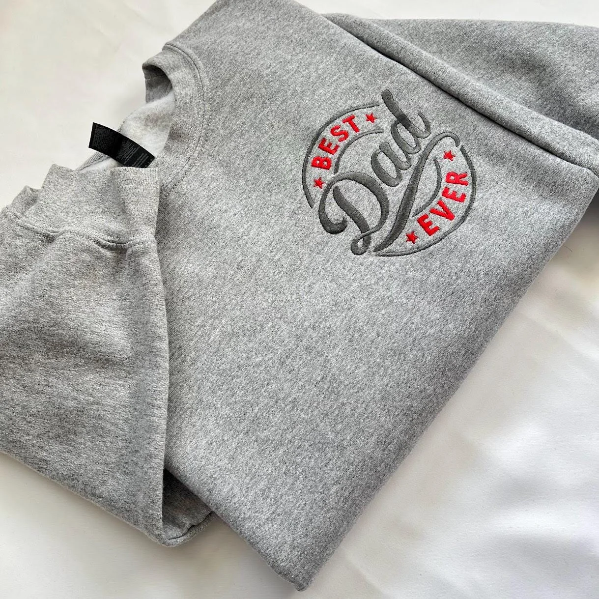 Angled view of a circular BEST Dad EVER badge with red stars and dark grey script stitched on a grey crewneck