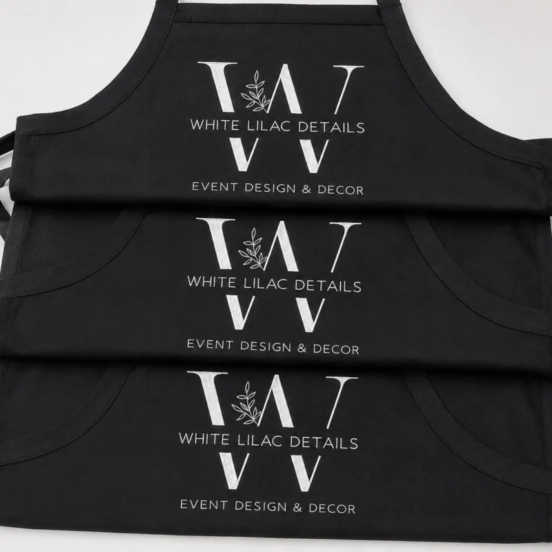 Three black aprons stacked, each with a white W monogram logo with a leaf sprig and an event business name