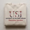 Sand crewneck with school initials in red and navy floral letters, the university name and a major in red script