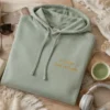 Sage crop hoodie flat-lay with gold Custom Text or Logo embroidery beside a matcha cup and headphones