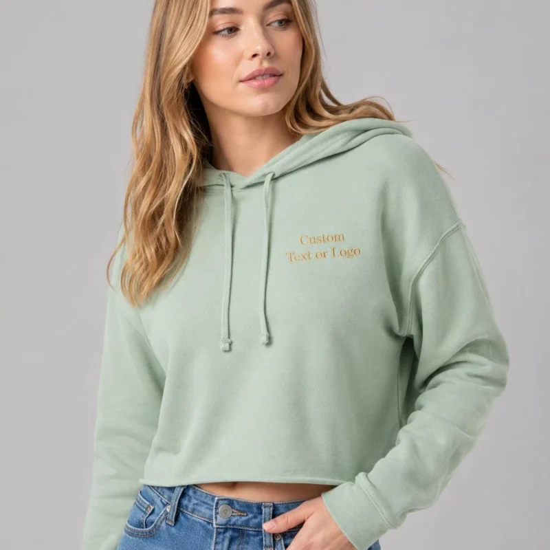 Woman in a sage crop hoodie and jeans with sample text Custom Text or Logo embroidered in gold on the left chest