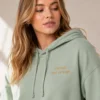 Woman in a sage cropped hoodie with small gold placeholder text on the chest, in soft window light
