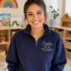 Smiling educator in a playroom wearing a navy quarter-zip with Sophia, ECE and Early Childhood Educator stitched