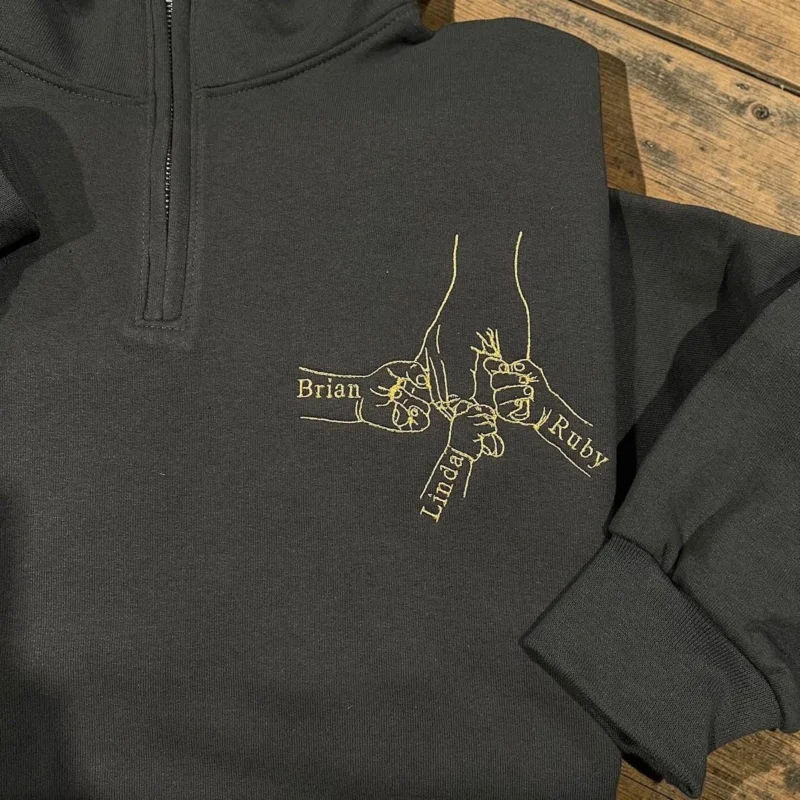 Close-up of gold outline hands meeting in a fist bump with the names Brian, Linda and Ruby on a charcoal quarter-zip
