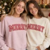 Two women in blush and bone crewnecks, each with MERRY in red gingham applique letters, by a Christmas tree