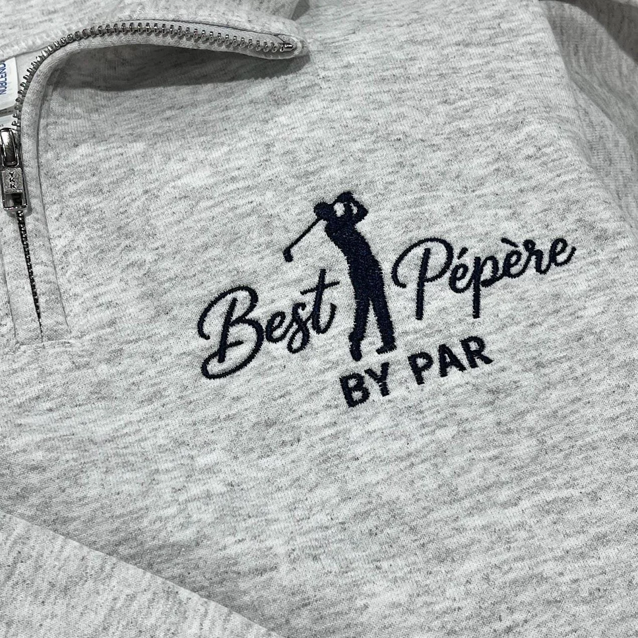 Close-up of a heather grey quarter-zip with the Best Pépère By Par golfer design in navy thread by the zip