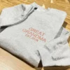 Angled ash grey crewneck with GREAT GRANDMA EST.2026 embroidered in coral thread, on a wooden table
