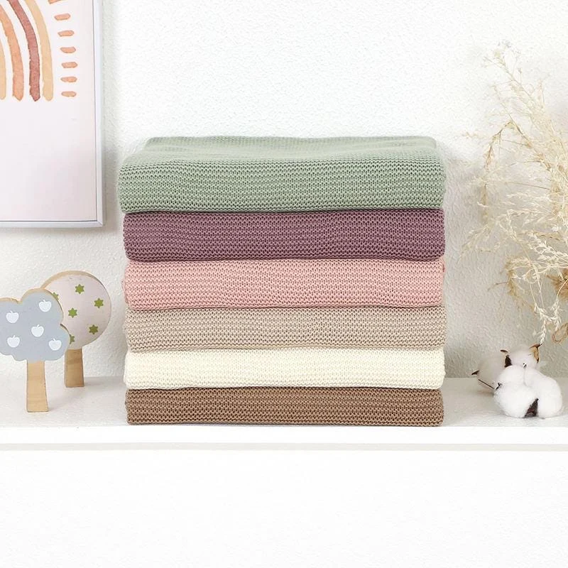 Stack of six folded knit baby blankets in sage, mauve, pink, khaki, ivory and taupe on a white shelf