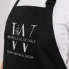 Angled close-up of a black apron bib showing the embroidered W monogram logo and EVENT DESIGN & DECOR