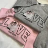 Pink and grey matching sweatshirts overlapped, each with LOVE in outline letters and an embroidered couple