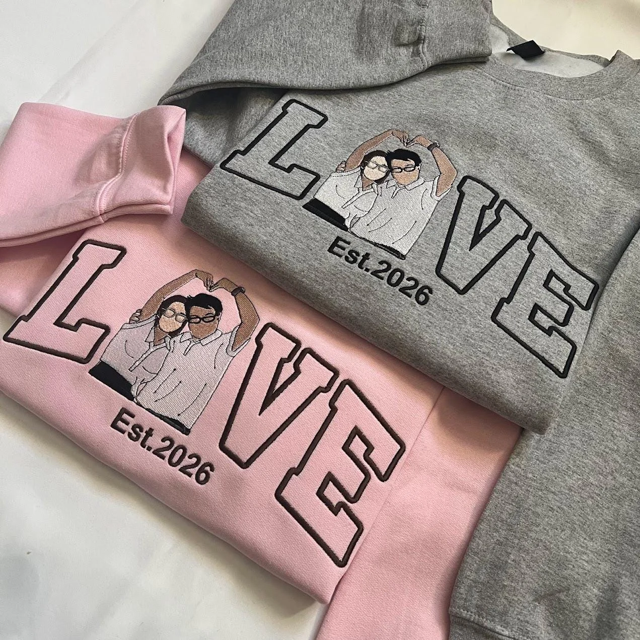 Pink and grey matching sweatshirts overlapped, each with LOVE in outline letters and an embroidered couple