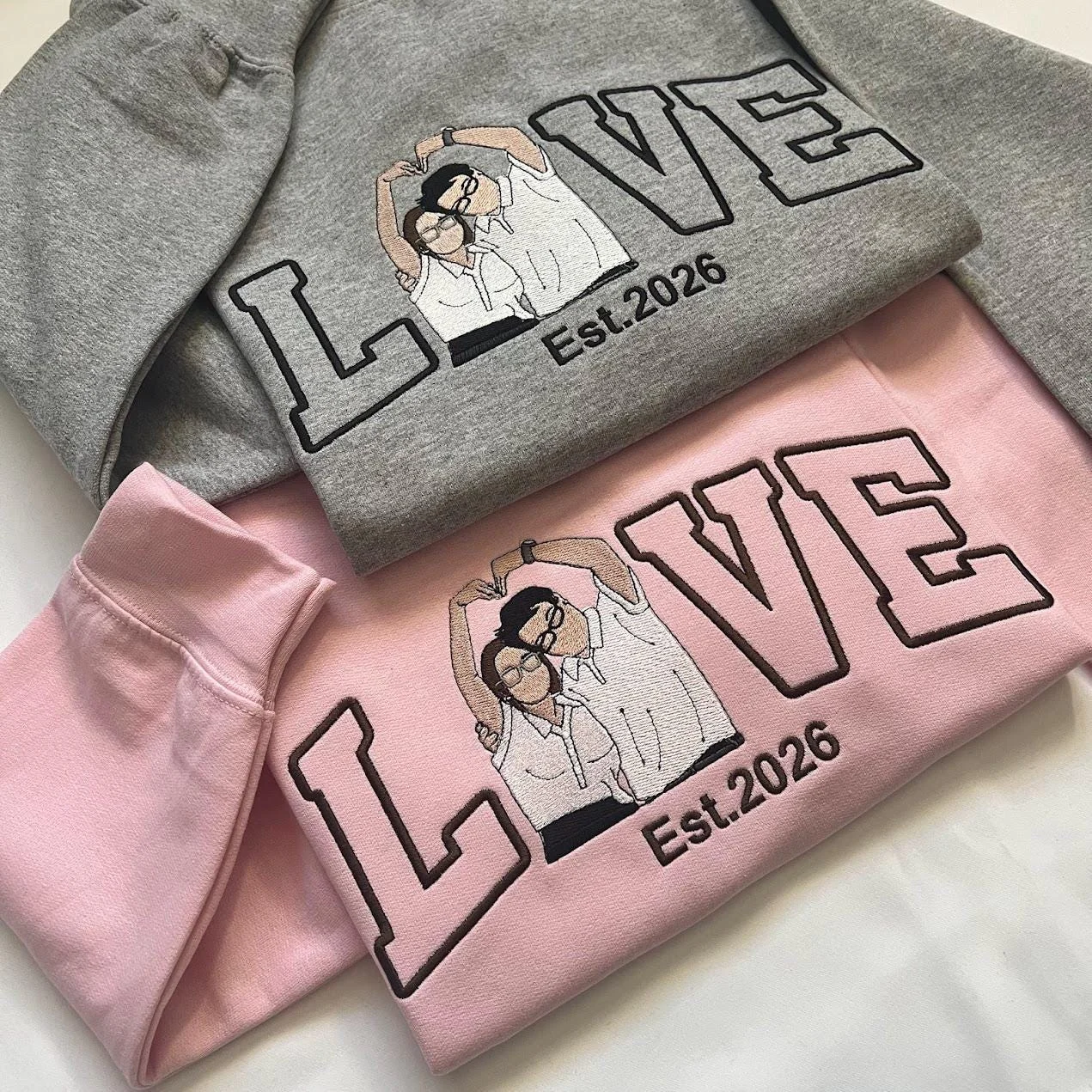 Angled grey and pink sweatshirts showing the stitched couple portrait inside LOVE lettering above Est.2026