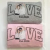 Folded grey sweatshirt stacked on a pink one, both with a couple portrait forming the O in LOVE and Est.2026