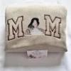 Folded sand sweatshirt with brown outline MOM letters and a stitched portrait of a mom cuddling her baby