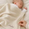 Sleeping newborn under an ivory knit blanket with Amelia stitched in mauve script at the lower corner