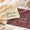 Ivory knit baby blanket with Mikail Carter in navy script and a mauve blanket with Faith in pink script, on a crib