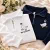 Matching white and navy quarter-zips with silly goose embroidery laid among roses, a ring box and a card
