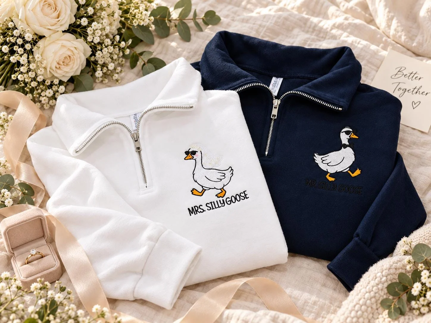 Matching white and navy quarter-zips with silly goose embroidery laid among roses, a ring box and a card